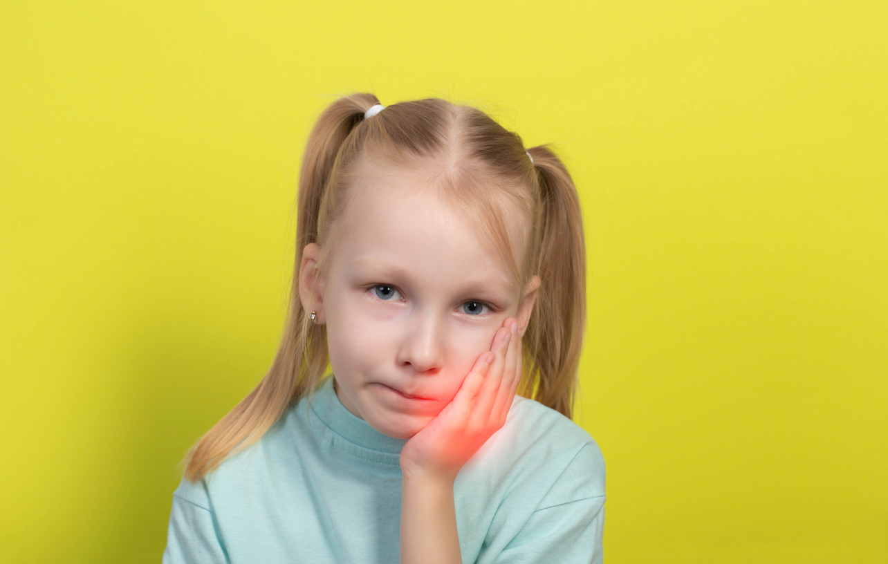 Young girl in front of a yellow background holding one hand against her jaw, which is illuminated by a red glow, because of cavity tooth pain.