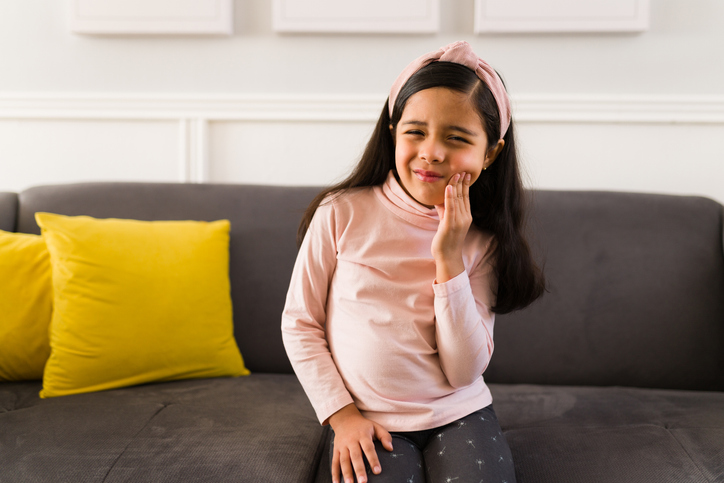 Young girl sitting on a couch rubbing her cheek in pain due to tooth sensitivity.
